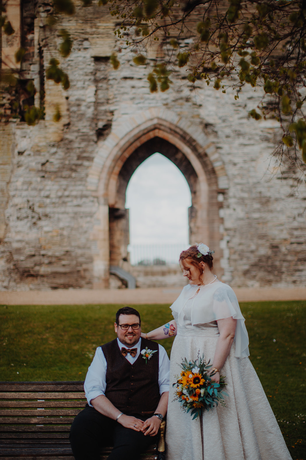 A man in a tweed waistcoat sits on a bench in front of a stone archway, his bride in a silk wedding dress stands at his side, looking down at him
